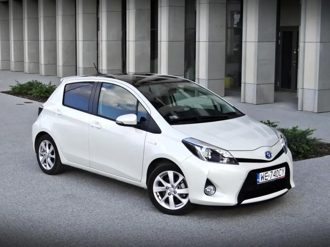 Biały Toyota Yaris z czarnym dachem, zaparkowany przed nowoczesnym budynkiem. Wiele osób szuka takich aut, czytając toyota yaris opinie.