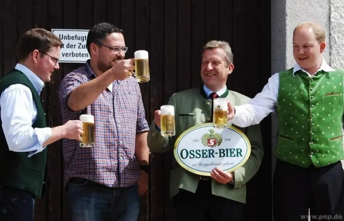 Vier Männer stoßen mit Biergläsern an. Einer hält ein Schild der Osser Brauerei.