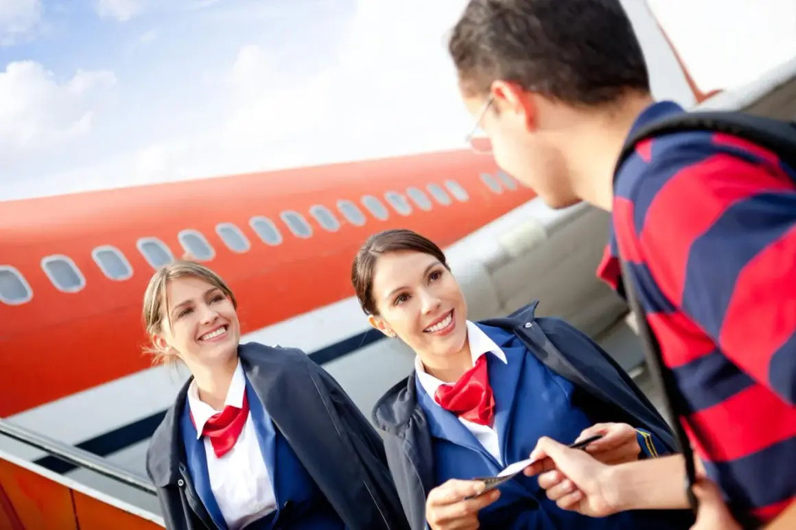 Zwei lächelnde Flugbegleiterinnen im blauen Anzug mit roten Halstüchern prüfen eine Bordkarte vor einem Flugzeug. Die Ausbildung zur Flugbegleiterin und das Gehalt sind attraktiv.