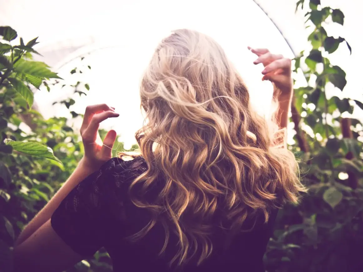 Frau mit welligem, blondem Haar, das durch Reverse Balayage einen natürlichen Farbverlauf erhält. Vorher-Nachher-Effekt im Grünen.