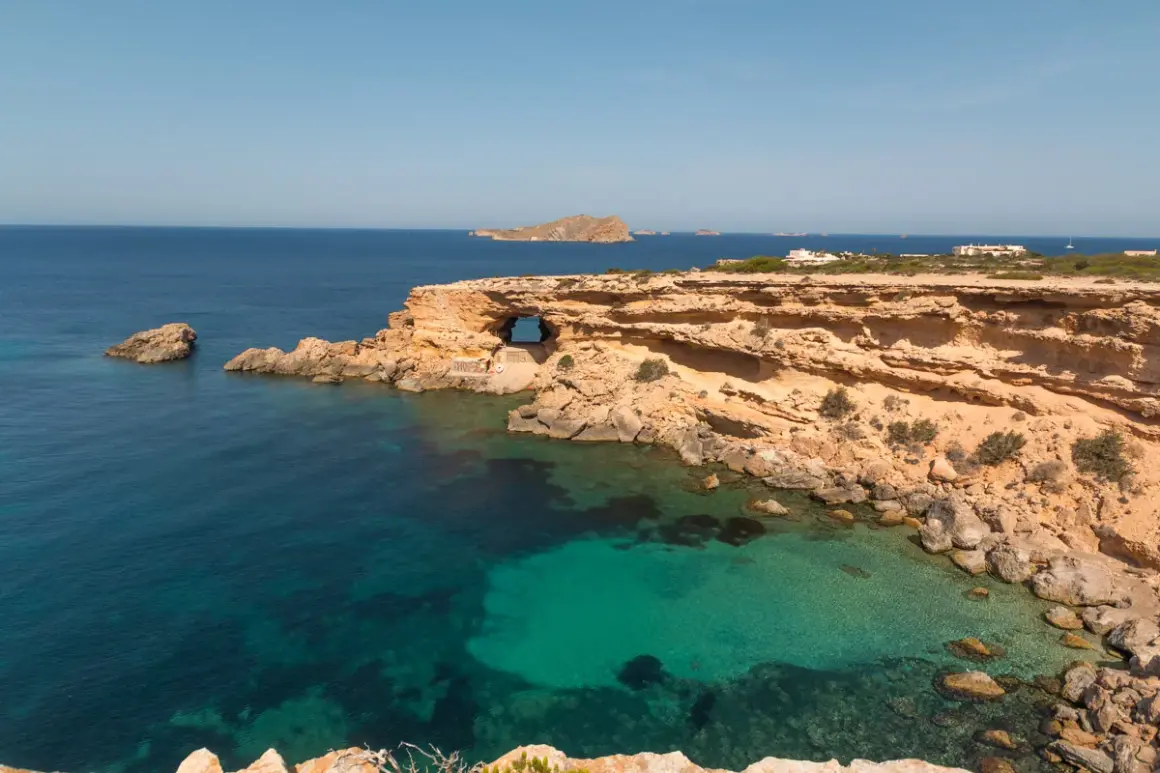 Costa rocosa con una cueva natural en Cala Comte, aguas turquesas y el mar azul profundo.
