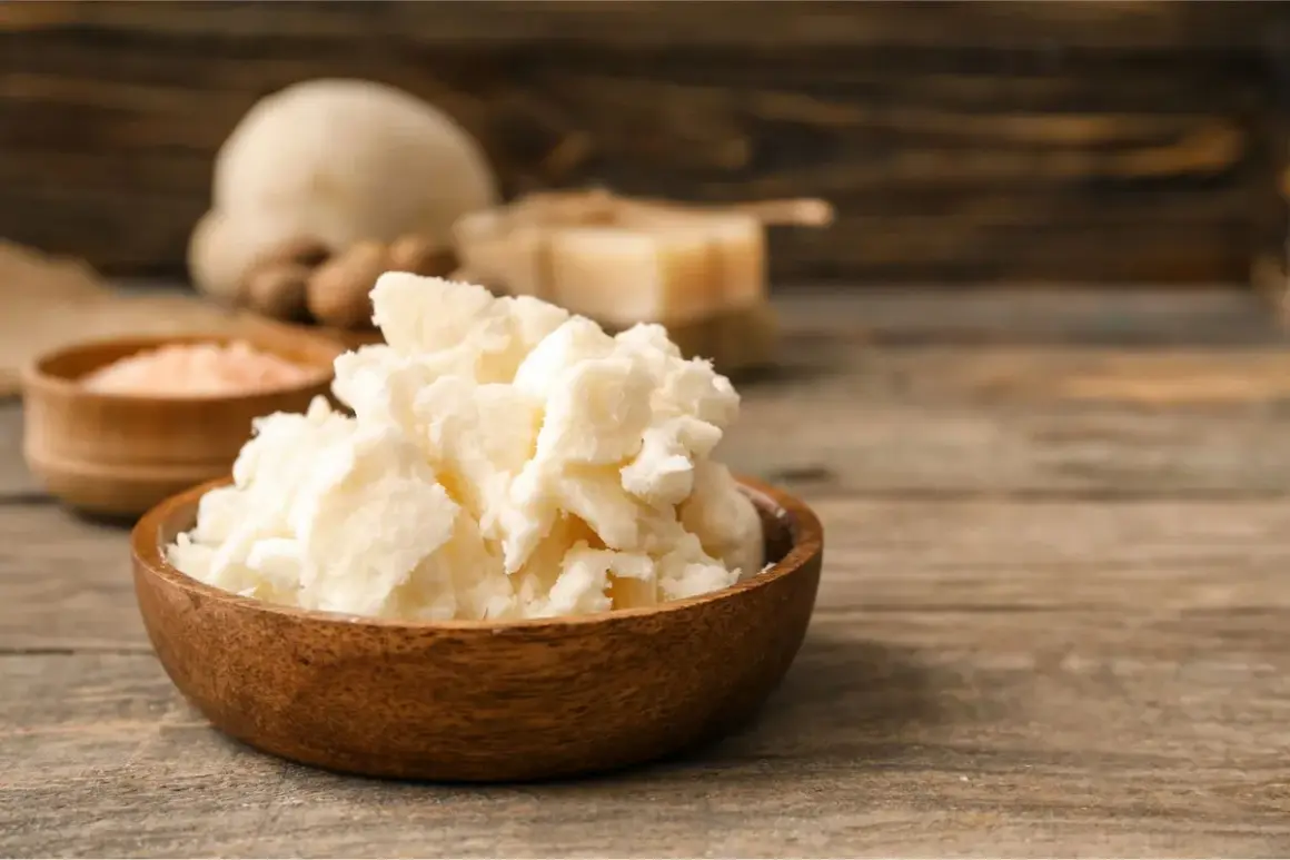 Un bol en bois rempli de beurre de karité, un ingrédient naturel pour les soins de la peau. Des noix de karité et du sel rose sont visibles en arrière-plan.