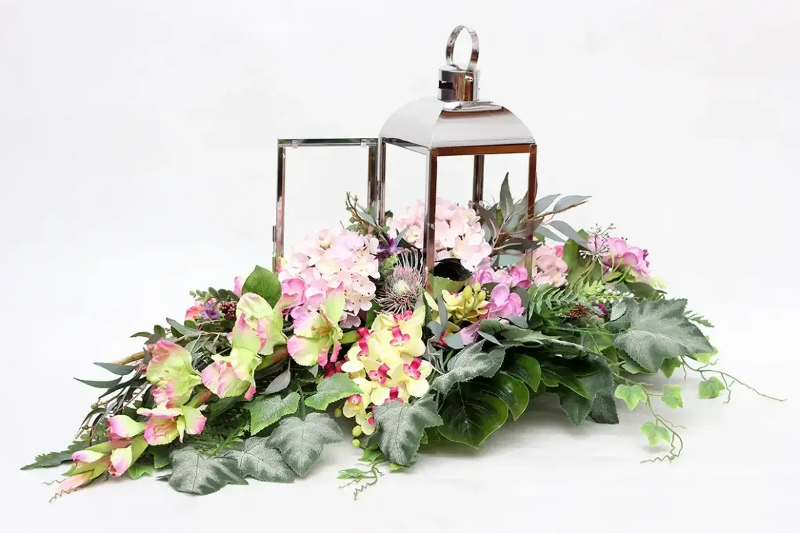 Elegancka kompozycja kwiatowa w lampionie, pełna różowych hortensji, żółtych i zielonych kwiatów, ozdobiona liśćmi i gałązkami.