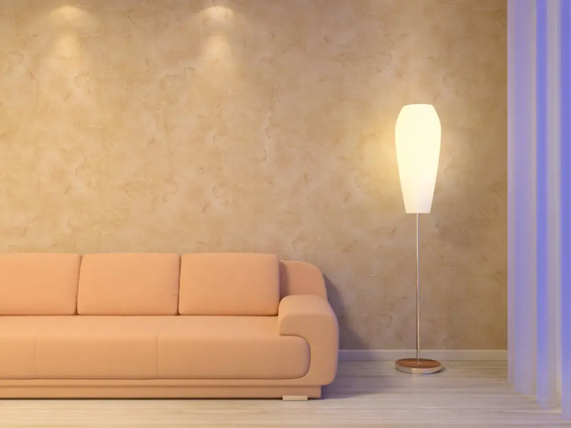 Miękka, brzoskwiniowa kanapa i elegancka lampa stojąca przy ścianie z fakturą przypominającą paca wenecka.