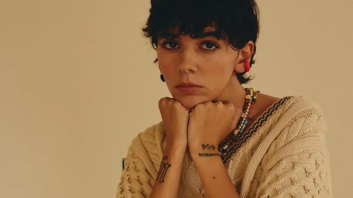 Un'ariete cantante con piercing e tatuaggi, indossa un maglione beige e una collana colorata.