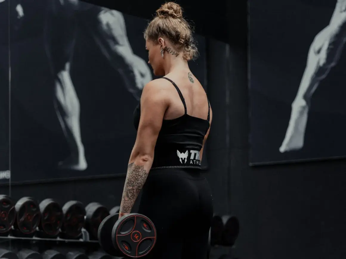 Kvinna med tatueringar utför shrugs med hantlar i ett gym.
