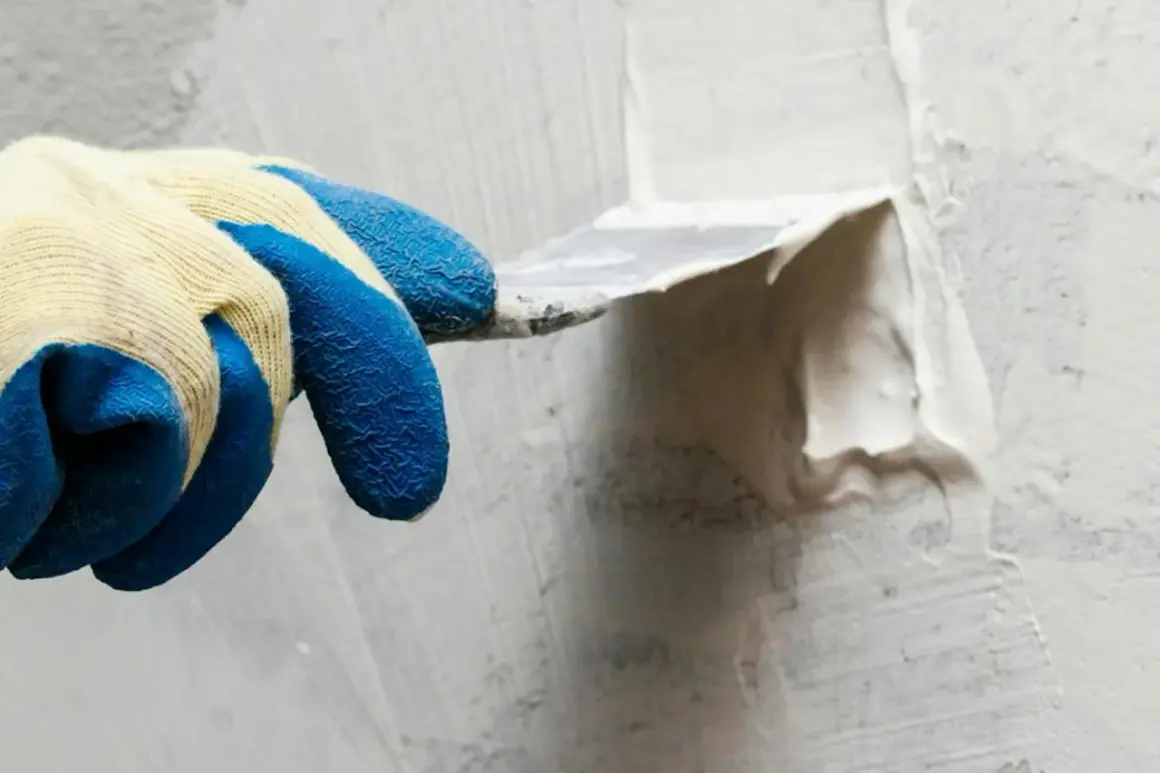 Hand med blå gummihandske spacklar en vägg utomhus. Snickerispackel utomhus används för att jämna till ytan.