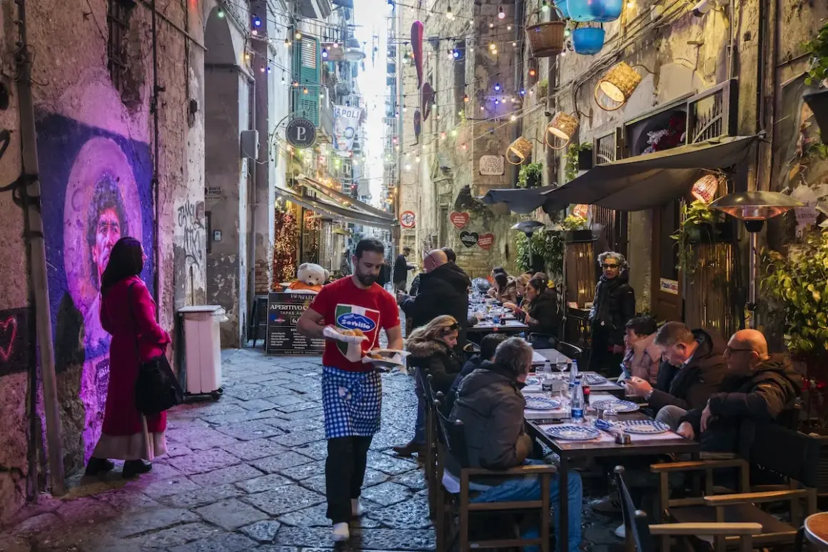Un vicolo napoletano illuminato da luci colorate, con gente seduta ai tavoli di un locale. Un cameriere porta del cibo.