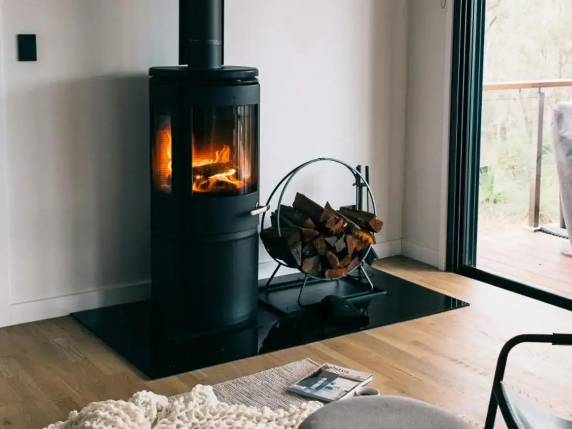 Moderner Holzofen mit brennendem Feuer, daneben ein Korb mit Holzscheiten. Ein gemütlicher Anblick, der an ein Verbot von Holzöfen denken lässt.