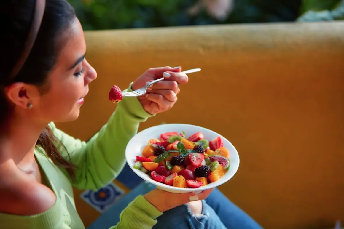 Une jeune femme savoure une salade de fruits colorée, une fraise à la fourchette. La naturopathie suggère quand manger les fruits pour une digestion optimale.