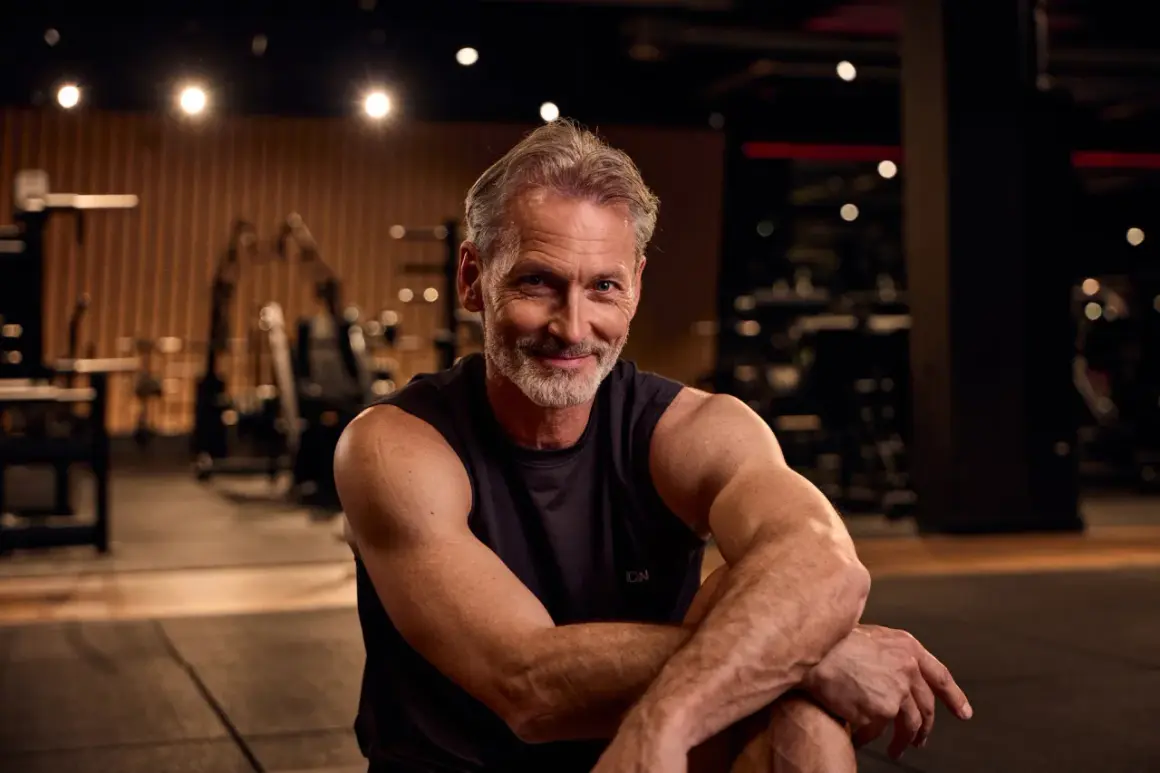 Ein lächelnder Mann mit grauem Haar und Bart sitzt im Fitnessstudio. Er zeigt, dass Muskelaufbau ab 50 möglich ist.