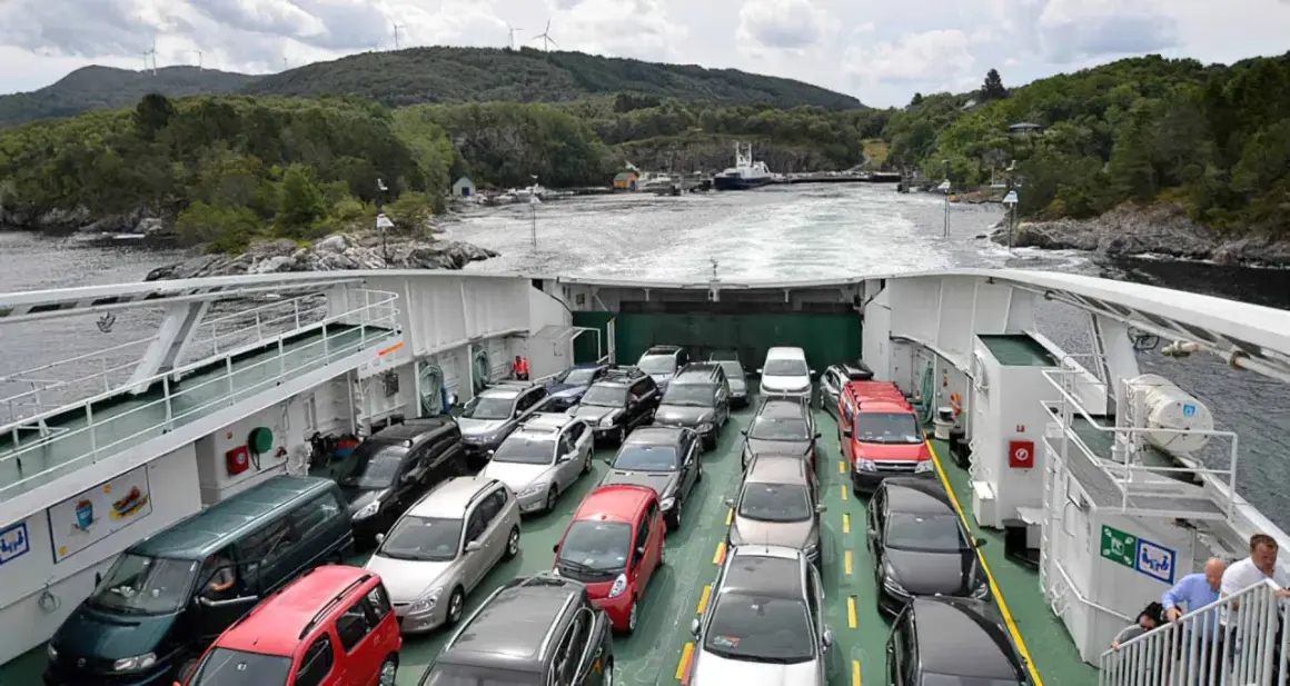 Coches en ferry cruzando un fiordo en Noruega. Paisaje verde y montañoso con molinos de viento.
