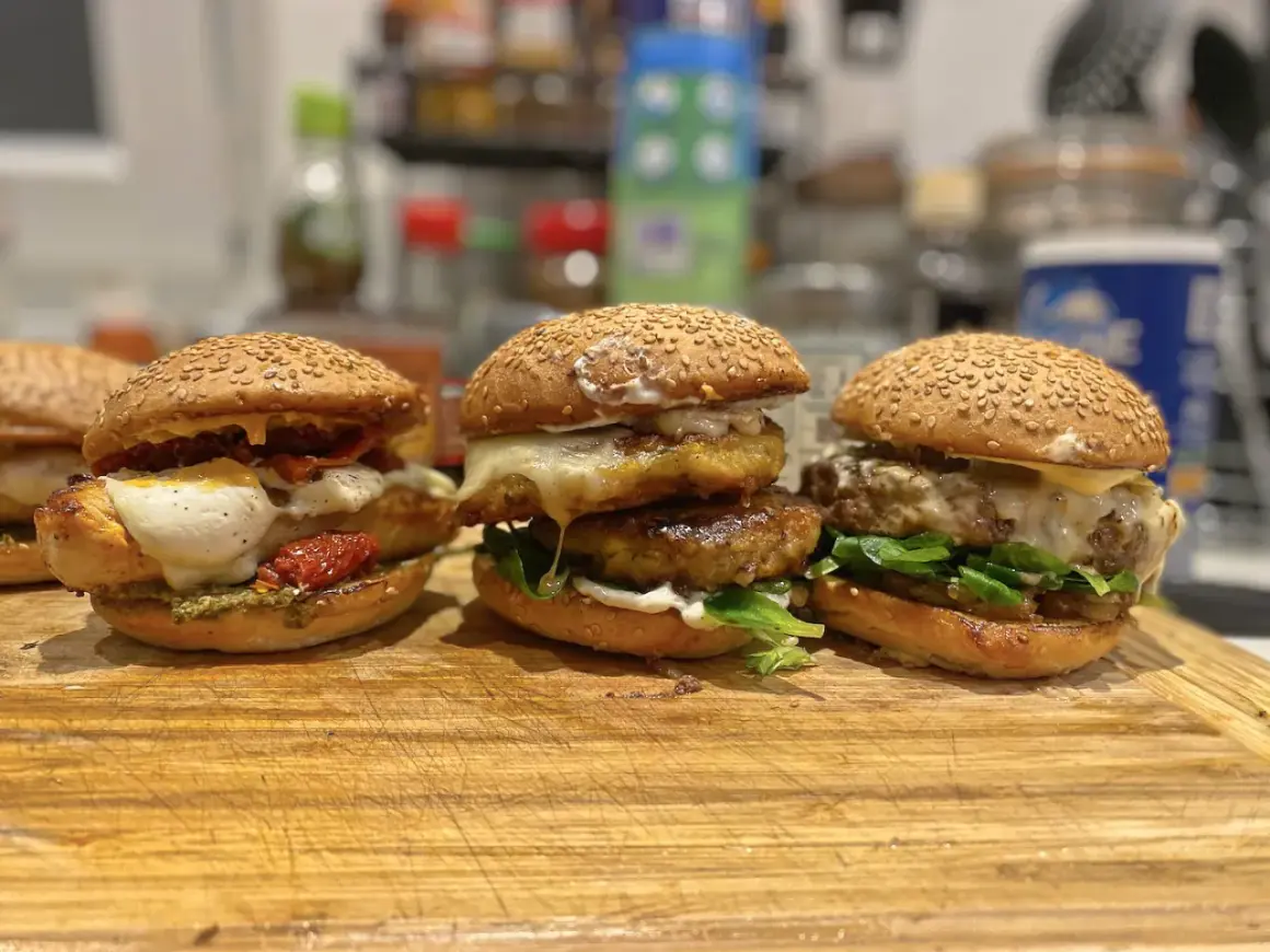 Trois burgers gras appétissants, garnis de fromage fondant, légumes frais et tomates séchées, prêts à être dégustés.