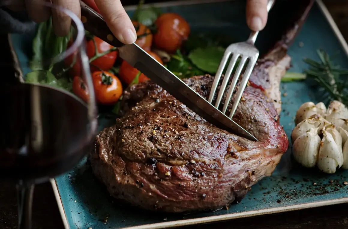 Ein saftiges Lamm-Steak wird mit Messer und Gabel angeschnitten. Dazu passen gerösteter Knoblauch, Kirschtomaten und ein Glas Rotwein. Welcher Wein zu Lamm? Ein kräftiger Rotwein!