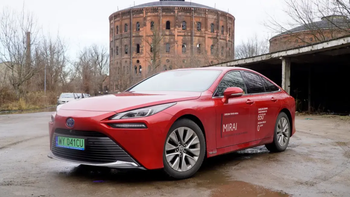 Czerwona Toyota Mirai z napisem "Zasięg na jednym tankowaniu do 650 km" stoi przed zabytkową gazownią.