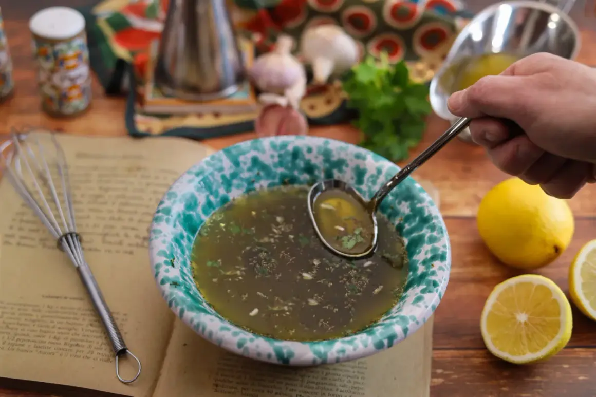 Un mestolo raccoglie il salmoriglio da una ciotola, pronto per la ricetta. Ingredienti freschi e un libro di cucina completano la scena.