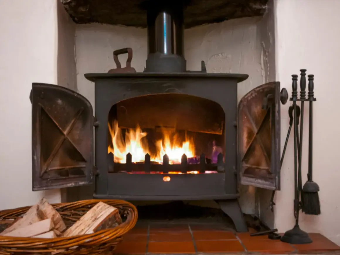 Ein Holzofen mit brennendem Feuer. Daneben ein Korb mit **Holz** und ein Werkzeugset. Die **Asche** glüht im Feuer.