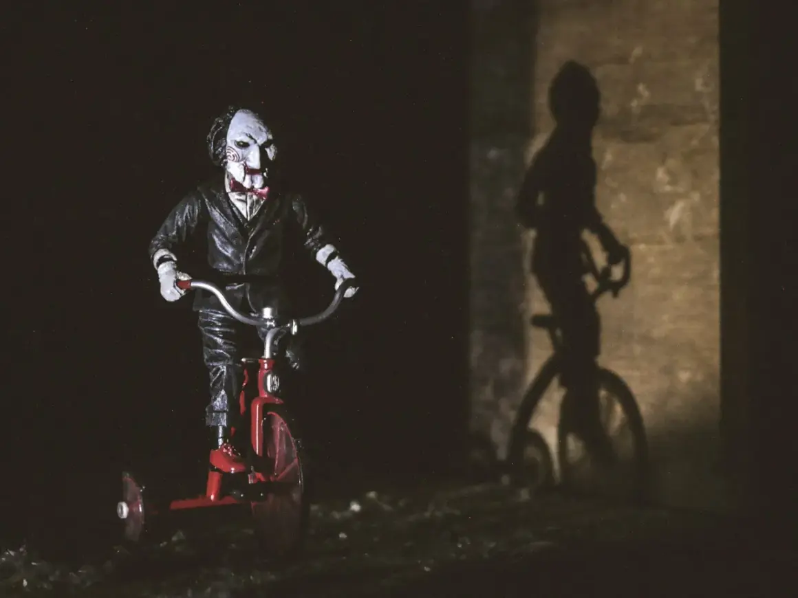 Lalka Billy z Piły na czerwonym trójkołowcu, cień na ścianie. Idealne do listy horrorów.