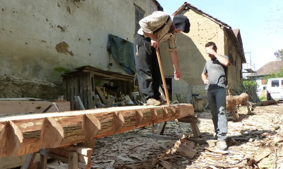Deux hommes travaillent sur un tronc d'arbre, pratiquant l'équarrissage bois avec une hache. Le bois est façonné avec soin.