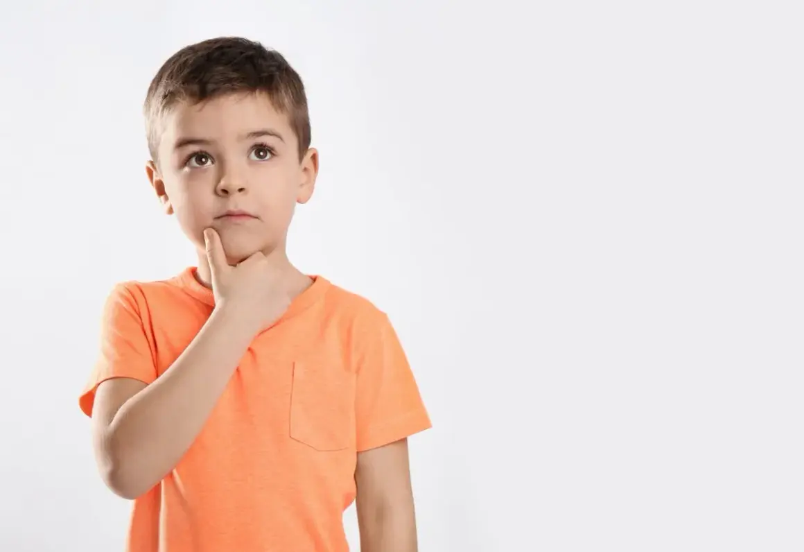 Jongetje met oranje shirt denkt diep na, misschien een beetje traag van begrip, met zijn vinger op zijn kin.