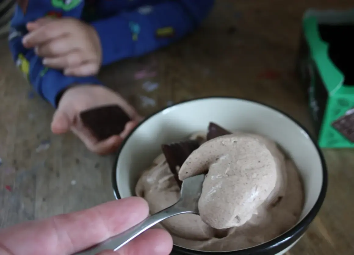 En sked med krämig after eight mousse lyfts ur en skål. En barnhand håller en chokladbit i bakgrunden.