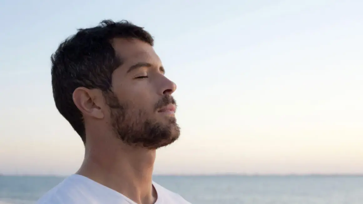Un homme au profil serein, yeux clos, savoure l'air marin. Sa barbe de 3 jours, d'environ 3 mm, accentue son charme naturel.