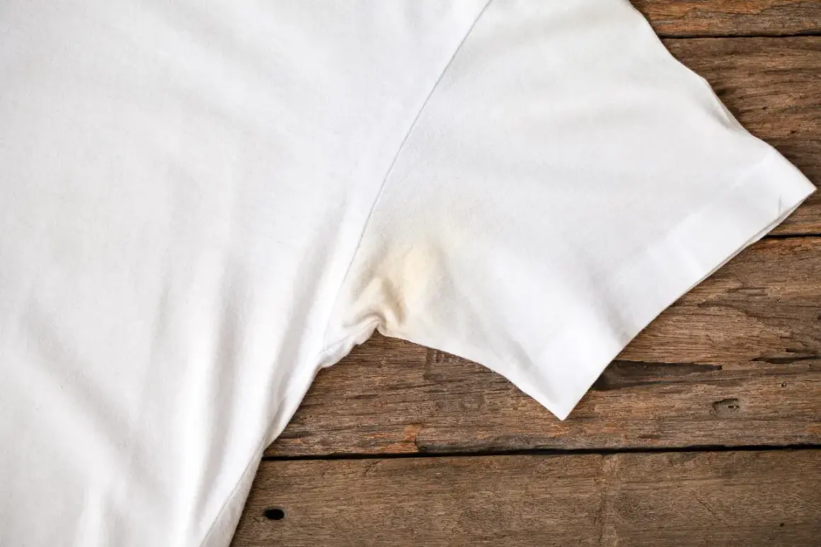 Verkrustete Deoflecken entfernen: Ein weißes T-Shirt mit gelblichen Flecken unter dem Arm auf Holzuntergrund.