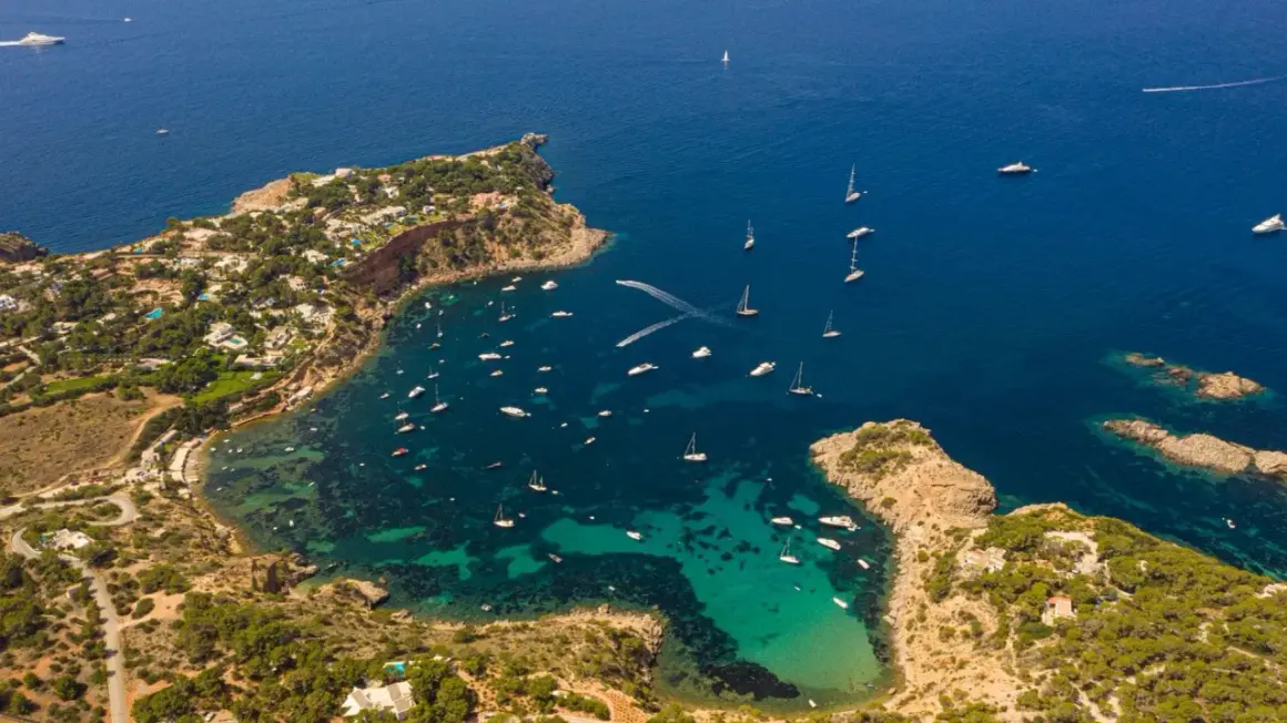 Una baia di Ibiza con numerose barche in fila, tra cui yacht e velieri, su acque cristalline.