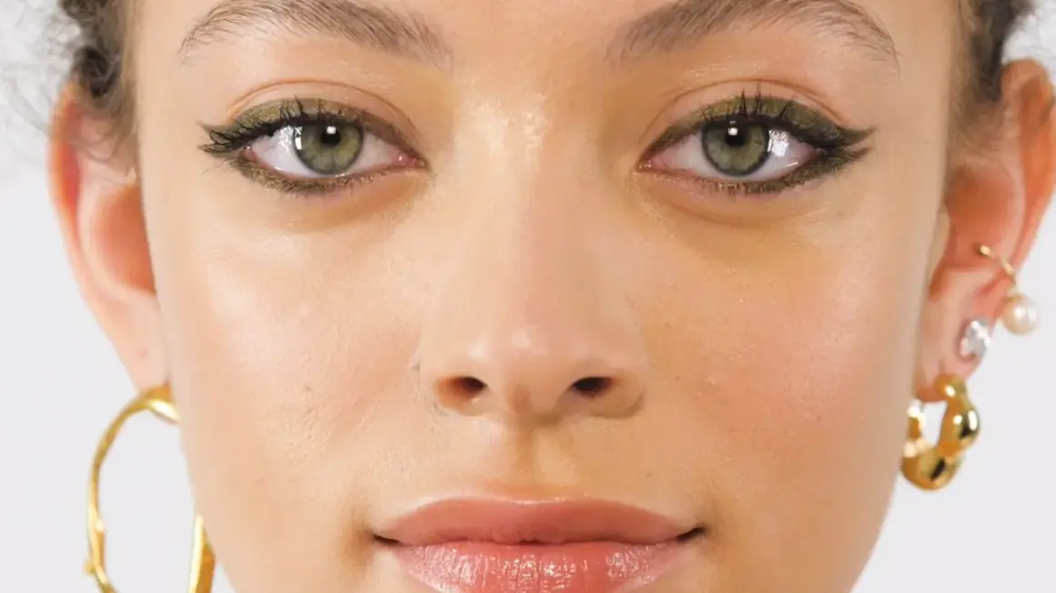 Primo piano di una ragazza con un trucco occhi audace e gioielli dorati. I suoi occhi verdi marroni sono evidenziati da un eyeliner scuro e scintillante.