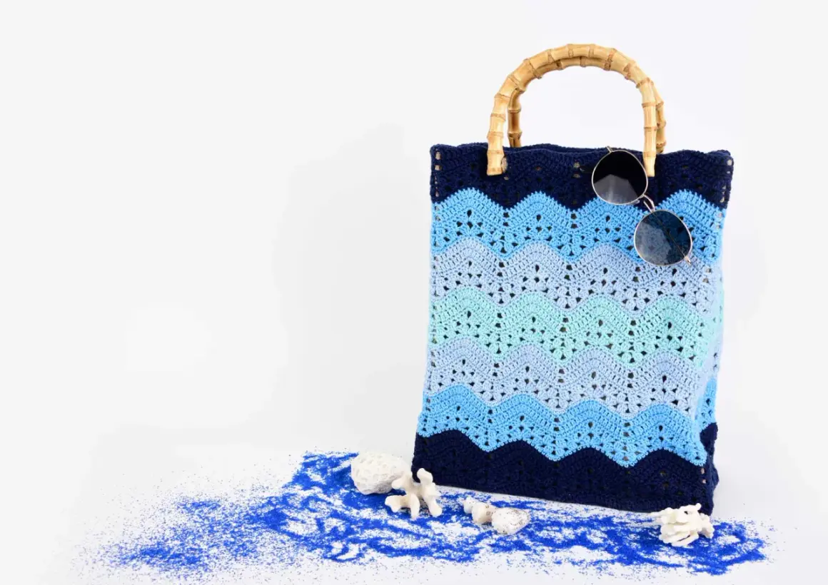 Borsa da spiaggia con manici in bambù, realizzata con splendidi lavori uncinetto cotone a onde blu. Occhiali da sole appoggiati sopra.