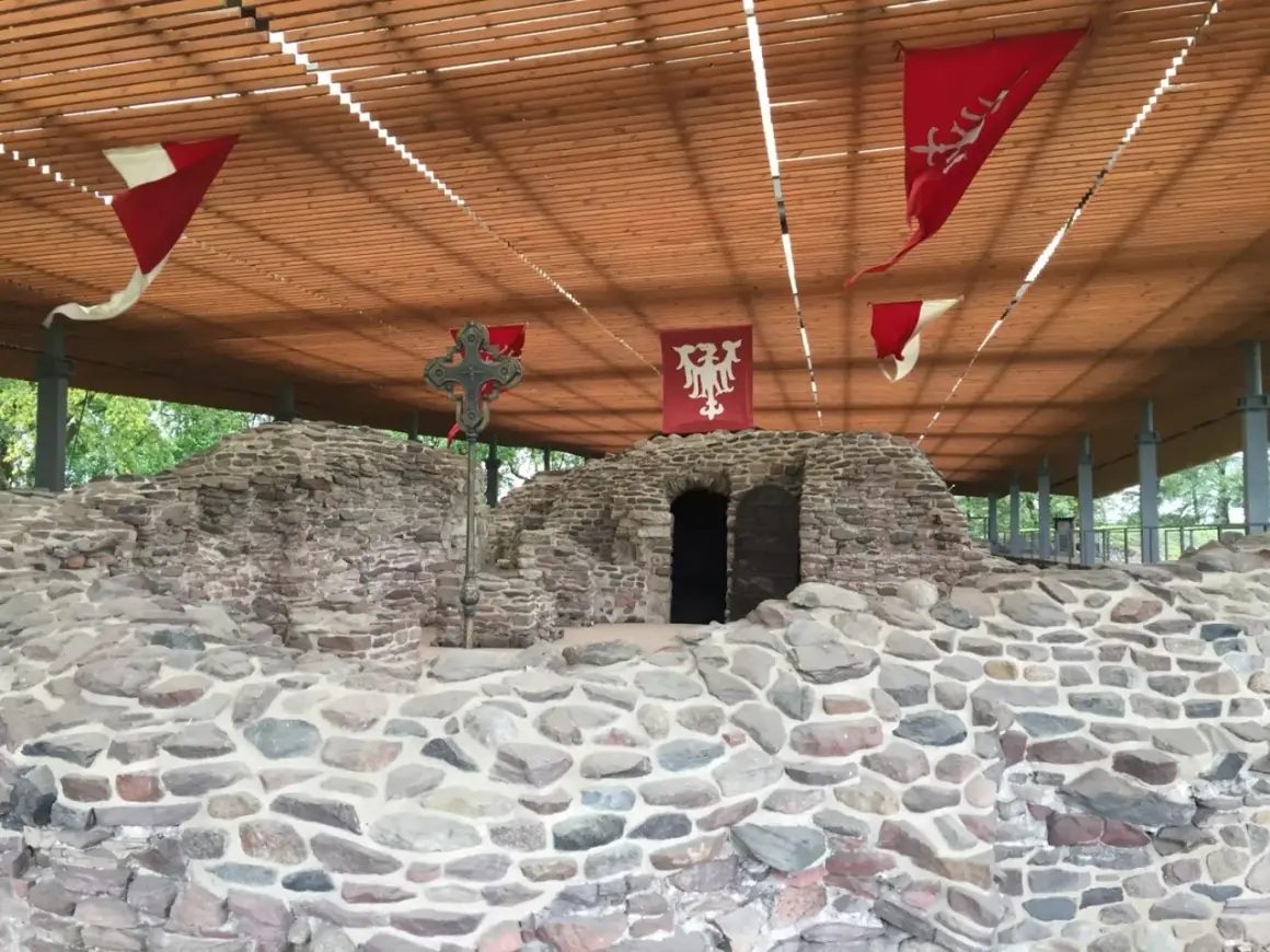 Kamienne ruiny na Ostrowiu Lednickim, z historycznymi chorągwiami i krzyżem. Wyobraź sobie, że to miejsce, skąd wyruszał dawny ostrów lednicki prom.