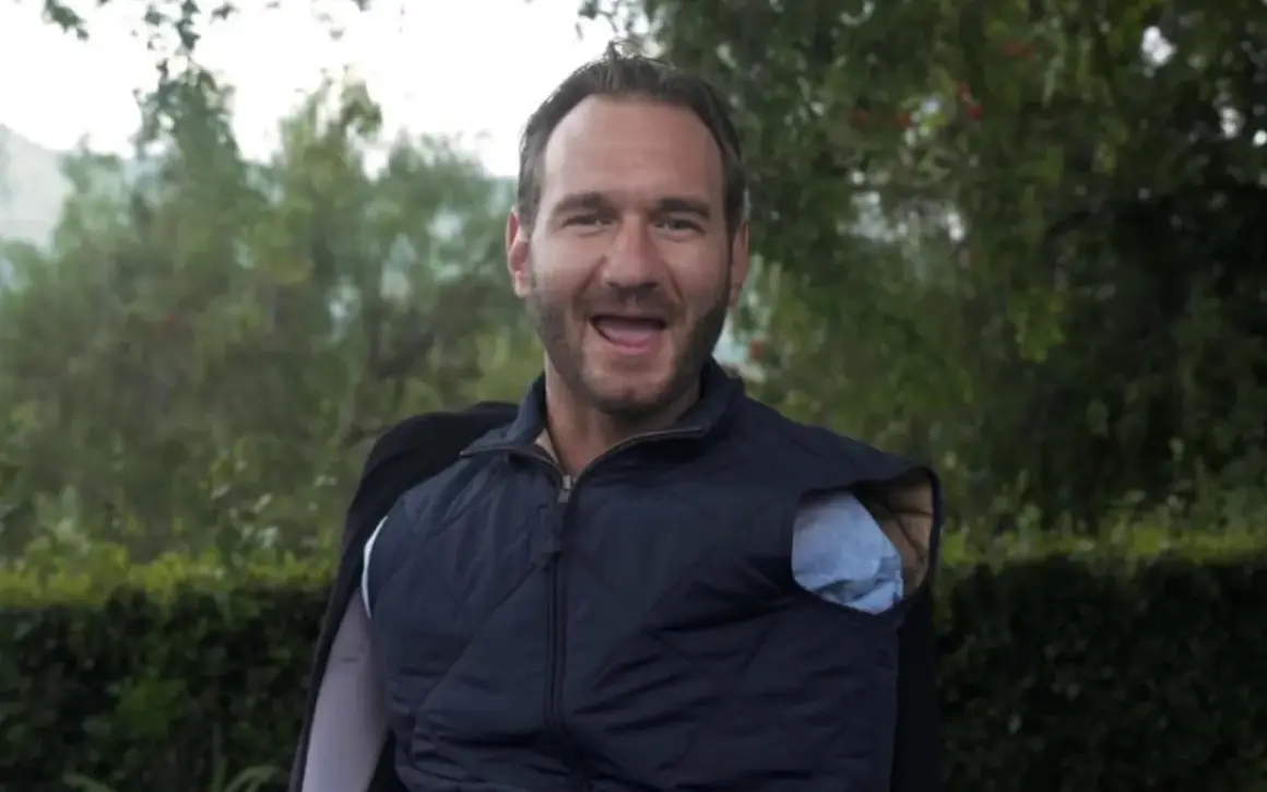 Nick Vujicic, lächelnd und mit offenem Mund, trägt eine blaue Weste über einem Hemd.