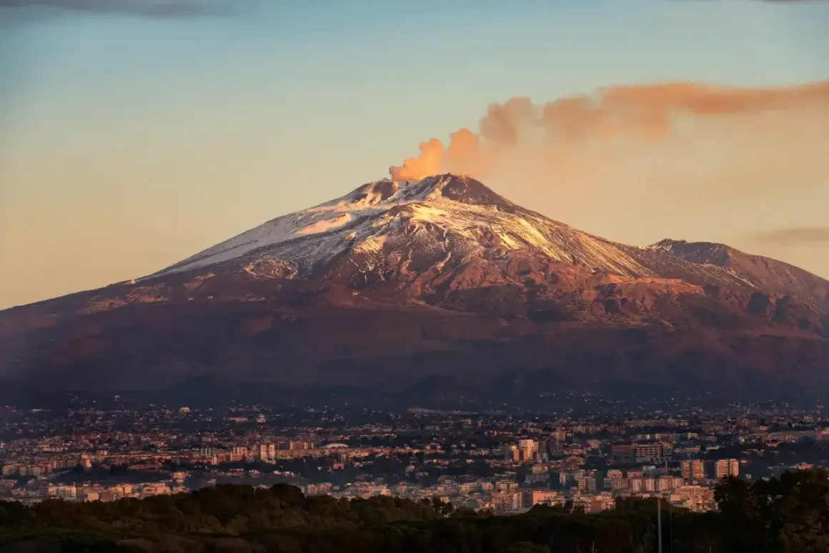 Etna, najwyższy wulkan Europy, dymi na tle zachodzącego słońca. U jego stóp rozciąga się miasto.