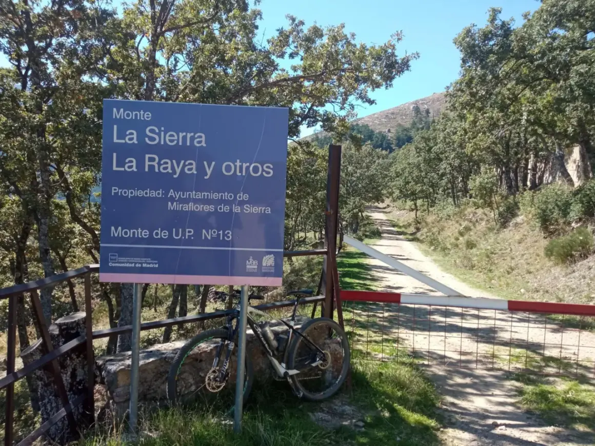 Señal de "Puerto de la Morcuera" junto a una bicicleta de montaña. Camino rural y árboles en un día soleado.