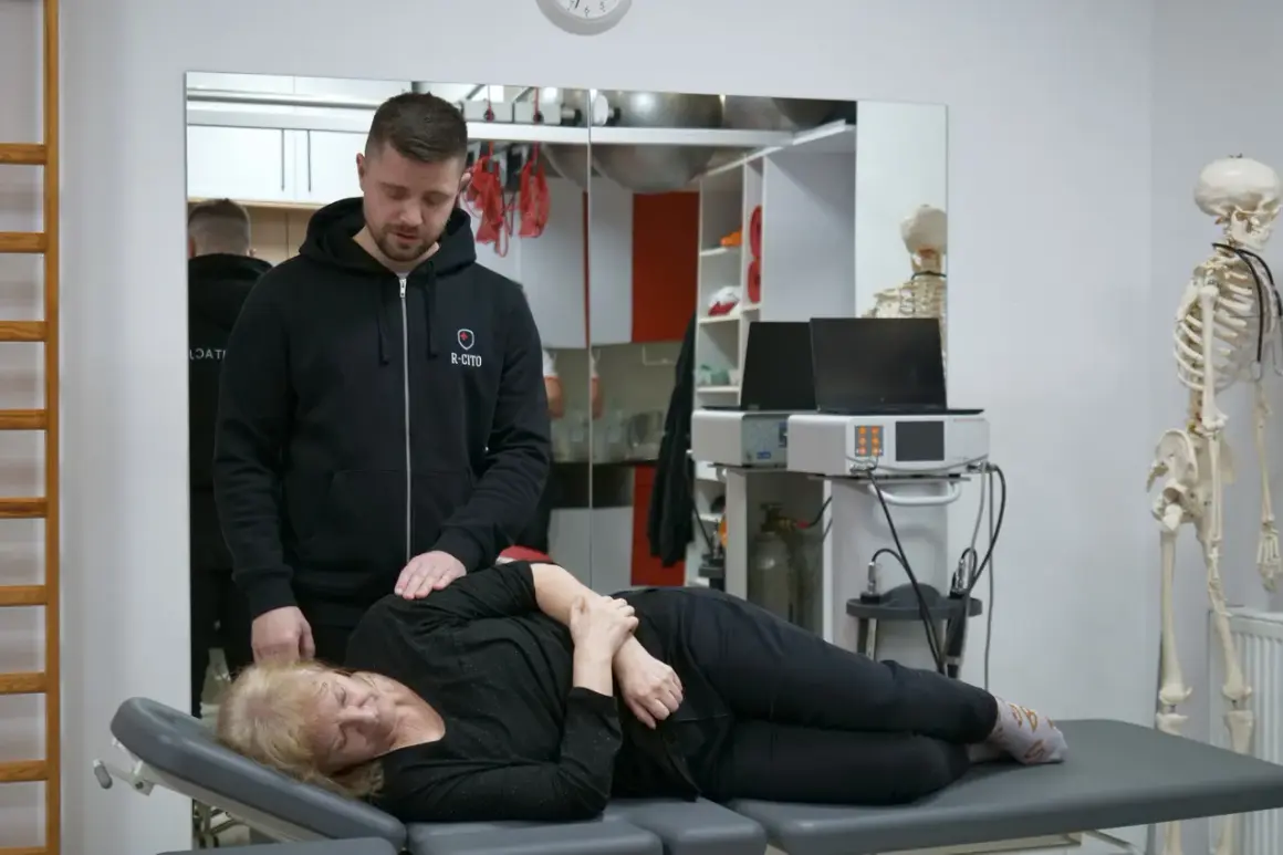 Fizjoterapeuta pomaga pacjentce, która cierpi na choroby od dźwigania. W tle model szkieletu i sprzęt rehabilitacyjny.