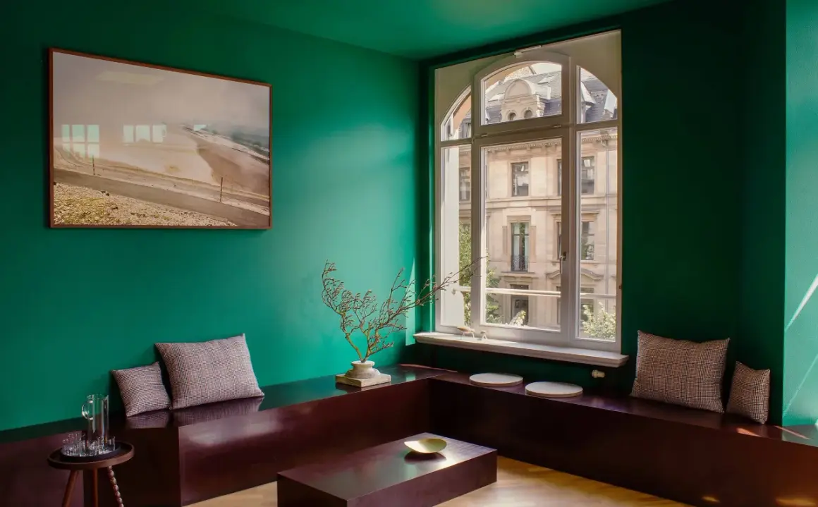 Un salotto con pareti verde scuro, un quadro paesaggistico, sedute in legno scuro e cuscini a quadri. I colori da abbinare al verde scuro includono il marrone del legno e le sfumature neutre dei cuscini.