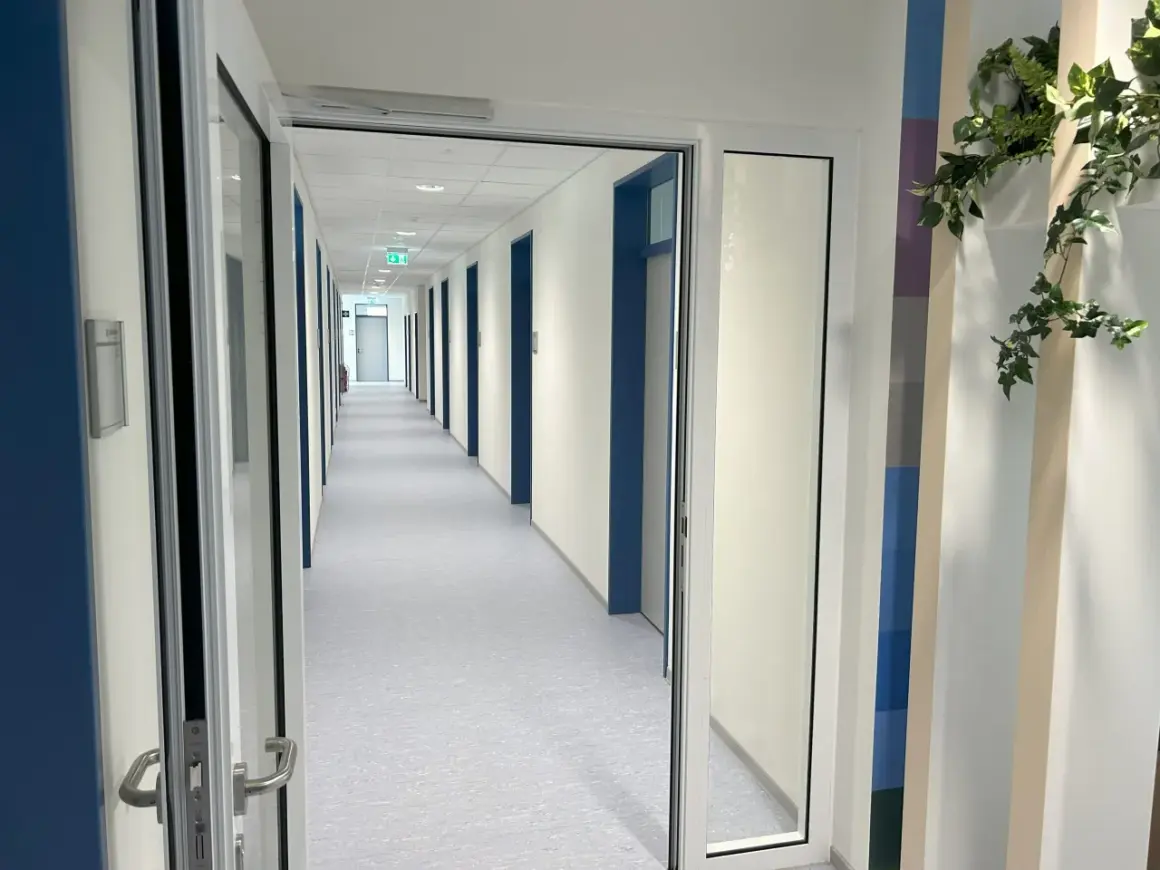 Langer Flur mit vielen Türen, beleuchtet von Deckenlampen. Grüne Notausgangsschilder sind sichtbar. Die Wartung von Feststellanlagen ist hier essenziell für die Sicherheit.