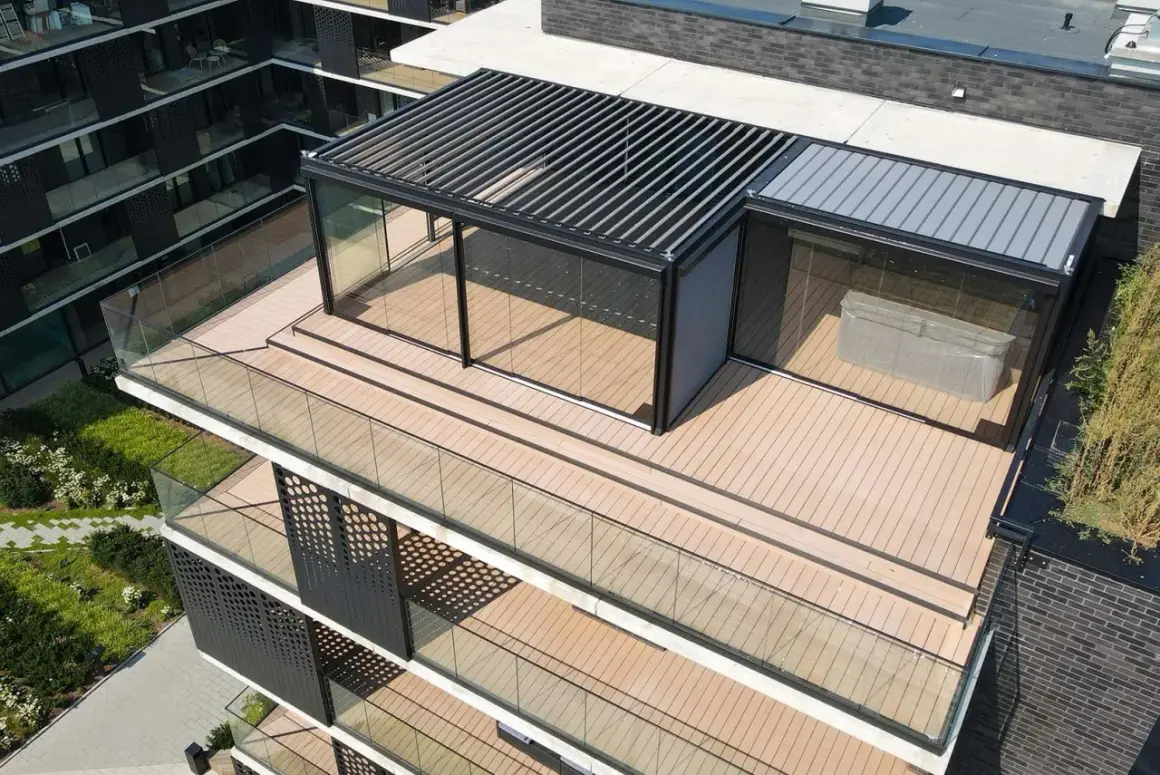 Nowoczesny taras na dachu z przeszklonym pawilonem i pergolą. Drewniana deska tarasowa i szklane balustrady tworzą elegancką przestrzeń.