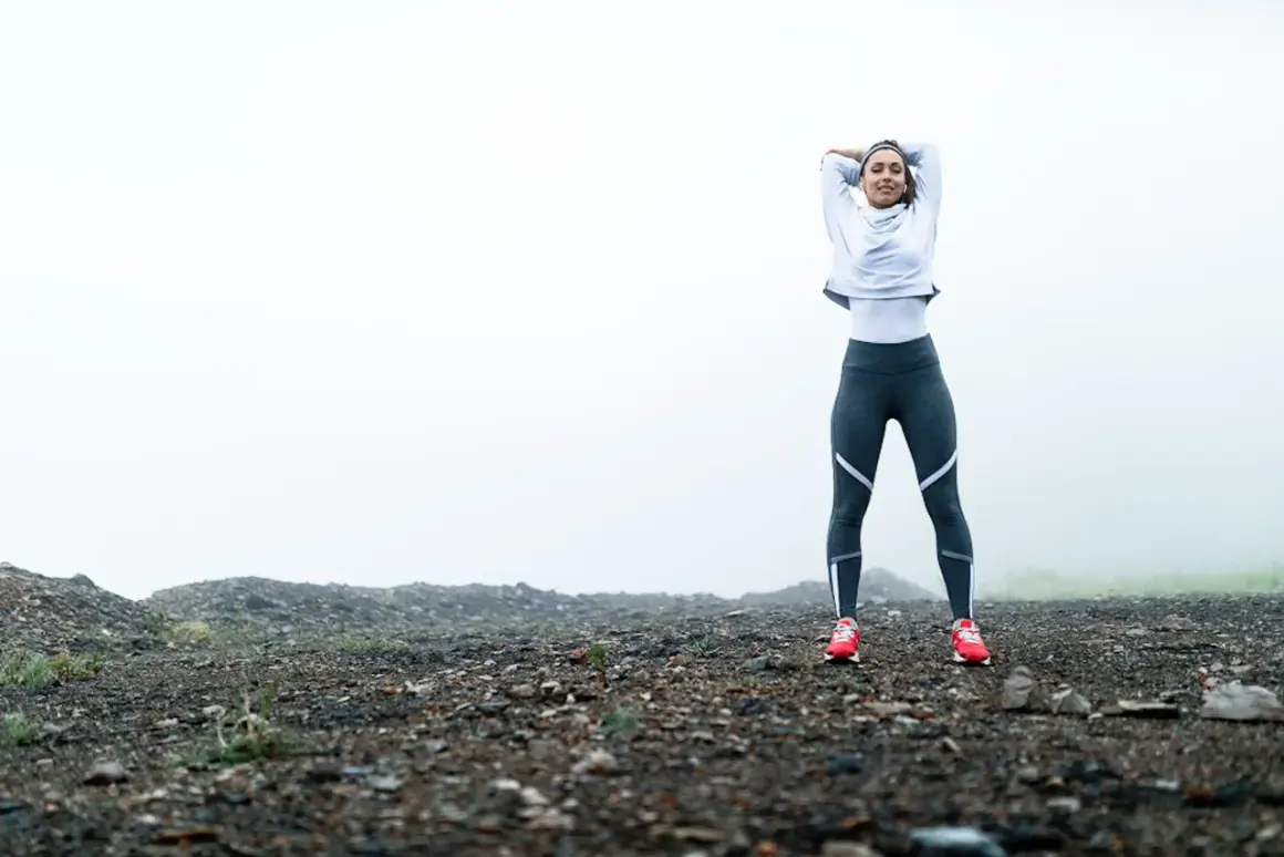 Femme en tenue de sport s'étirant sur un terrain rocailleux, prête pour son entrainement course à pied dans un paysage brumeux.