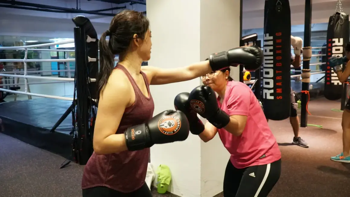 Two women spar in a boxing gym. One woman in a maroon tank top throws a punch, while the other in a pink shirt dodges. They both wear boxing gloves, ready for a cruiser weight bout.