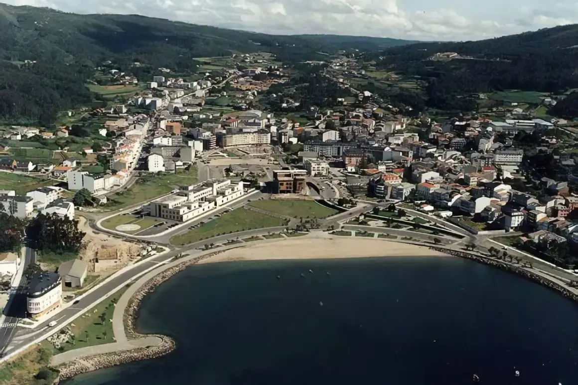 Vista aérea de la playa de Cee, con el mar azul, la arena dorada y la ciudad extendiéndose hacia las colinas verdes.