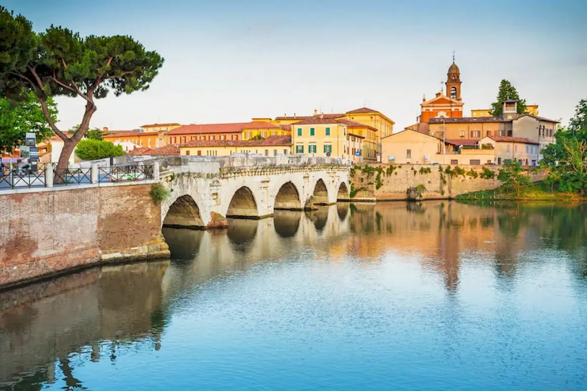 Il Ponte di Tiberio a Rimini, con i suoi archi in pietra che si specchiano sull'acqua calma, fa da sfondo a un paesaggio urbano colorato.