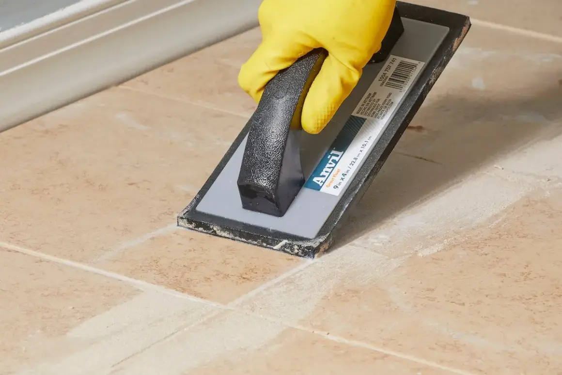 A gloved hand uses a grout float to spread grout, leaving a light grout haze on the tiled surface.