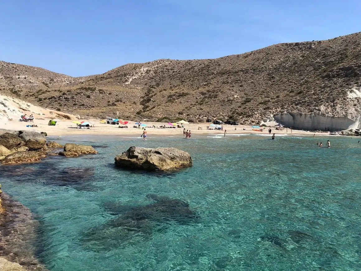 Aguas cristalinas en la cala del medio, con gente disfrutando del sol y el mar en un día soleado.