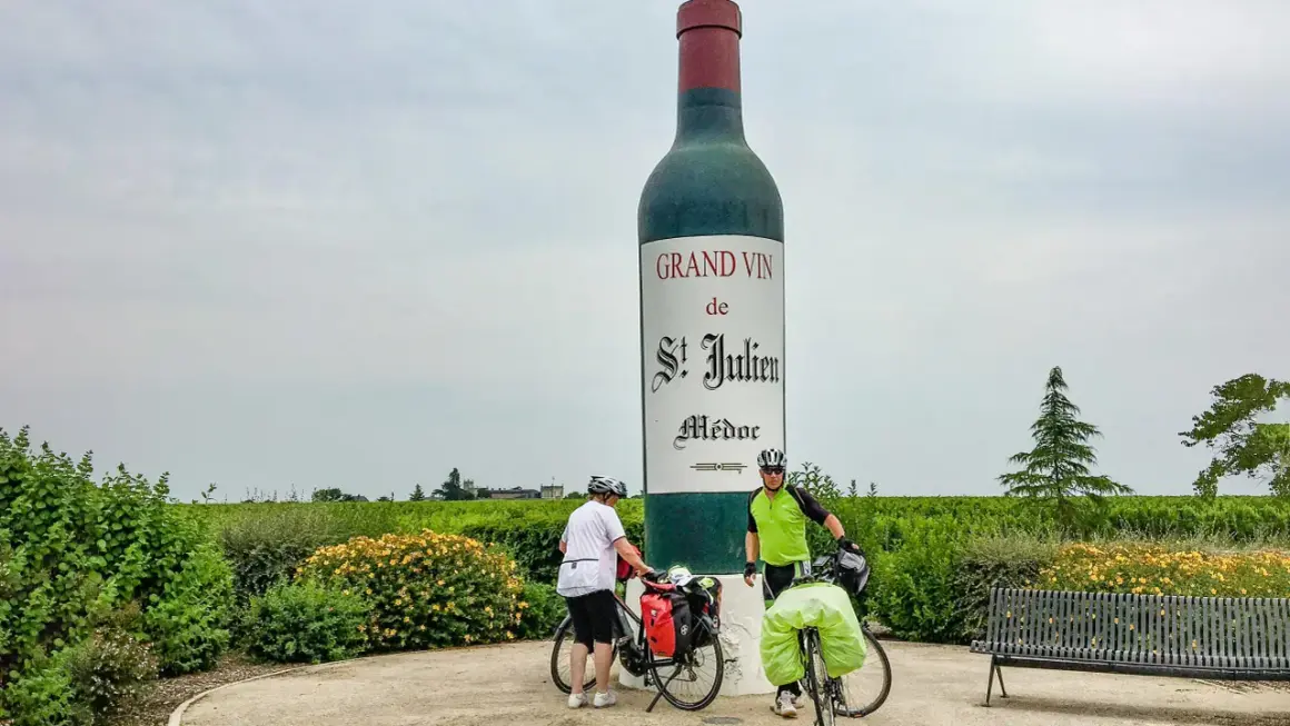 Zwei Radfahrer pausieren vor einer riesigen Weinflasche in Medoc, Frankreich. Weinberge erstrecken sich bis zum Horizont.