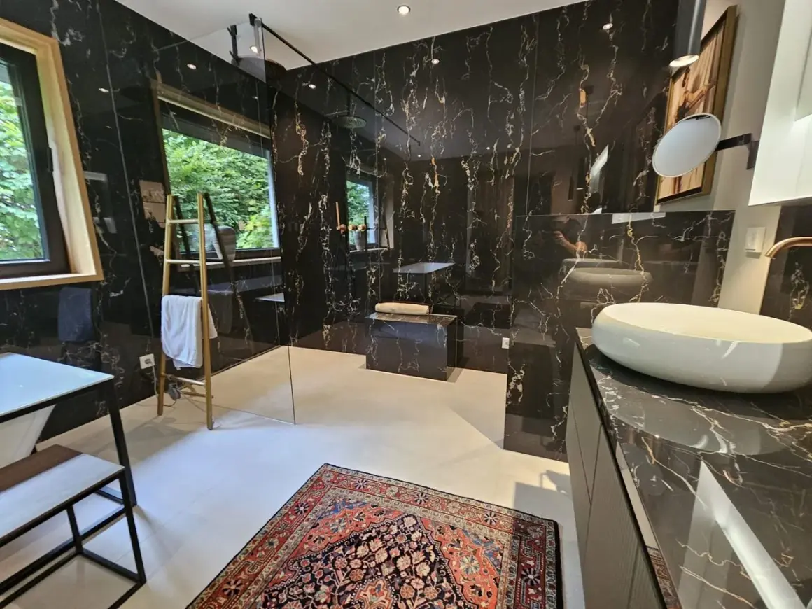 Luxuriöses Bad ohne Fenster mit schwarzem Marmor, freistehender Badewanne, Dusche und Waschbecken. Ein bunter Teppich liegt auf dem Boden.