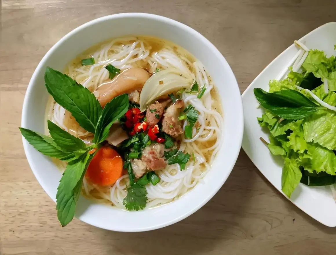 Aromaticzny makaron pho z kawałkami mięsa, warzywami i świeżymi ziołami. Obok talerz z sałatą i kiełkami fasoli.