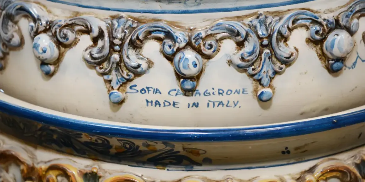 Dettaglio di un'opera in porcellana bianca e blu, con decorazioni barocche e la firma "Sofia Caltagirone Made in Italy".