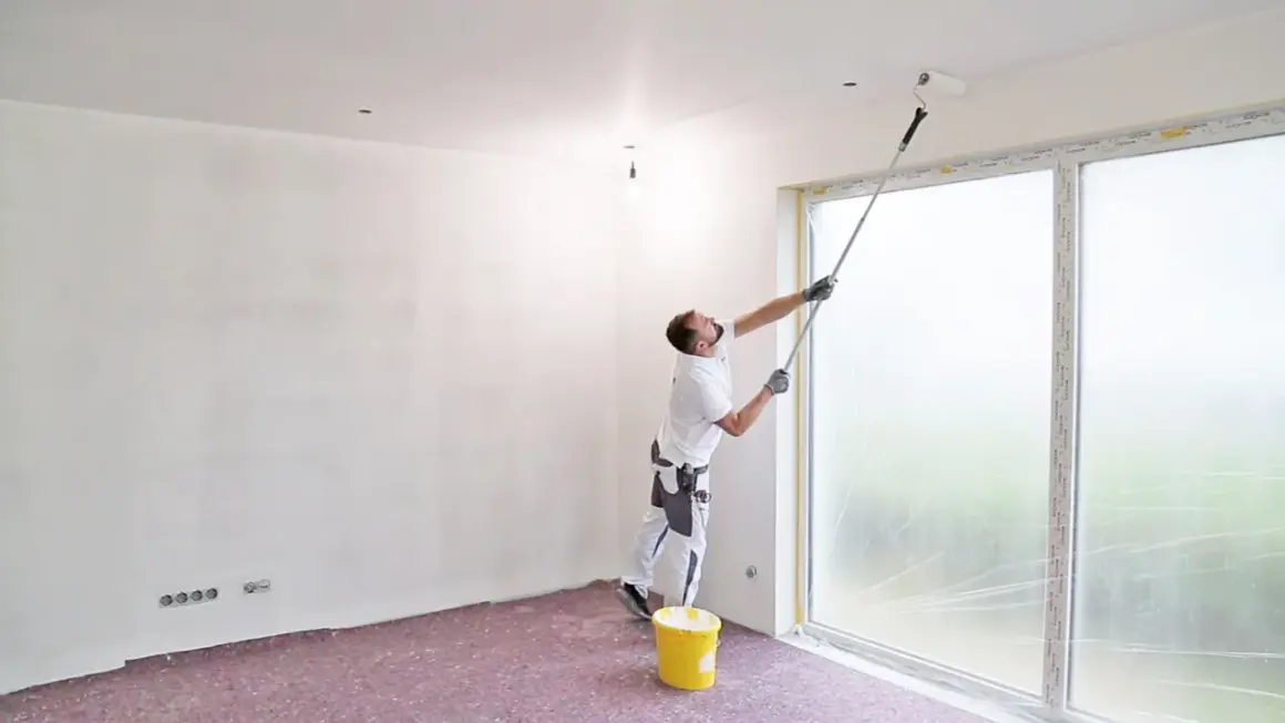 Maler streicht die Decke mit einer Teleskopstange. Ein gelber Farbeimer steht bereit.