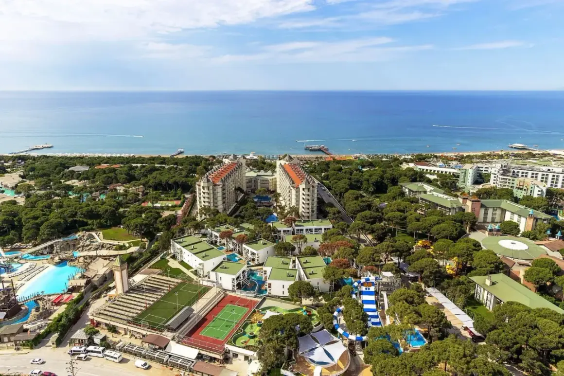 Widok z lotu ptaka na kompleks hotelowy z basenami, zjeżdżalniami i boiskami sportowymi, otoczony zielenią, nad turkusowym morzem. Idealne miejsce na wakacje w Turcji.